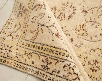 May include: A cream-coloured area rug with a folded corner revealing the underside. The rug features a repeating pattern of brown floral and geometric motifs, a wide decorative border with intricate linear details, and a short fringe edge.