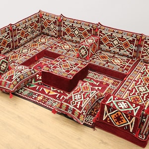 May include: A modular floor seating set in a rich burgundy color, featuring intricate geometric patterns in shades of cream, orange, green, and brown. The set includes multiple cushions and a central square ottoman, all adorned with tassels.