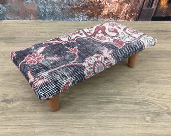 May include: A rectangular footstool with a patterned top in shades of navy, red, and cream. The design features floral motifs. The stool is supported by four short, light-brown wooden legs. The overall aesthetic is vintage or antique.