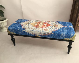 May include: A rectangular bench featuring a cushioned top upholstered in a vibrant blue fabric with a central medallion pattern in red, orange, and white. The bench is supported by four black, turned wooden legs with a smooth, glossy finish.