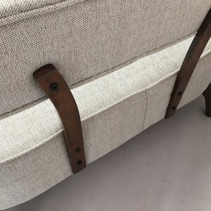 May include: A beige upholstered sofa featuring a textured fabric finish and curved dark brown wooden support legs attached to the backrest with visible black screws. The sofa has a structured frame with a clean, neutral-toned textile.