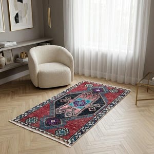 May include: A rectangular, vintage-style rug with a central geometric design in shades of red, blue, and black. The rug is placed on a light-colored wooden floor, with a cream-colored chair and a window with white curtains in the background.