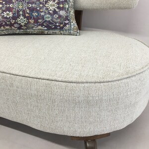 May include: A curved sofa upholstered in a light beige textured fabric with a dark wooden base. A rectangular throw pillow rests on the seat, featuring a deep purple background with a complex, multi-coloured floral and geometric pattern.