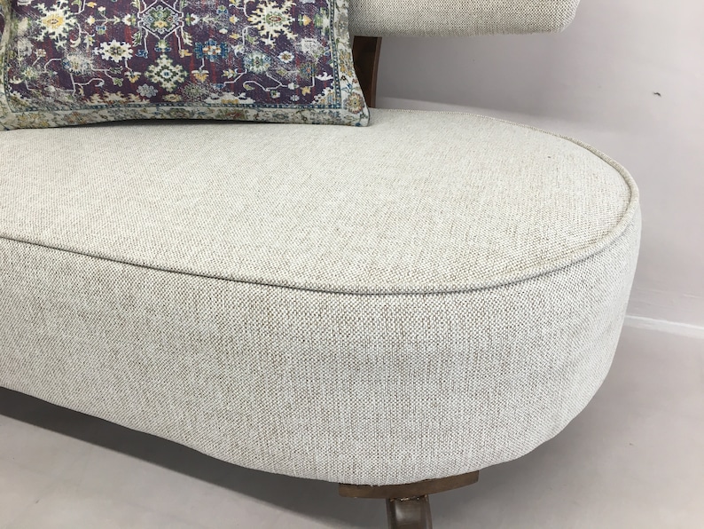 May include: A curved sofa upholstered in a light beige textured fabric with a dark wooden base. A rectangular throw pillow rests on the seat, featuring a deep purple background with a complex, multi-coloured floral and geometric pattern.