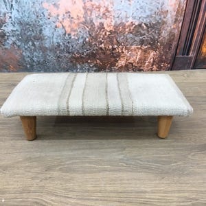 May include: A small, rectangular ottoman with a woven beige and brown fabric top and wooden legs. The ottoman is sitting on a wooden floor.