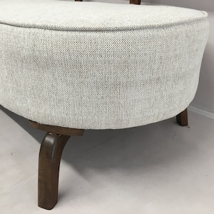 May include: A curved bench featuring light grey, textured fabric upholstery with a visible weave pattern. The seat is supported by dark brown wooden legs that curve outwards, providing a stable base for the rounded, upholstered furniture piece.
