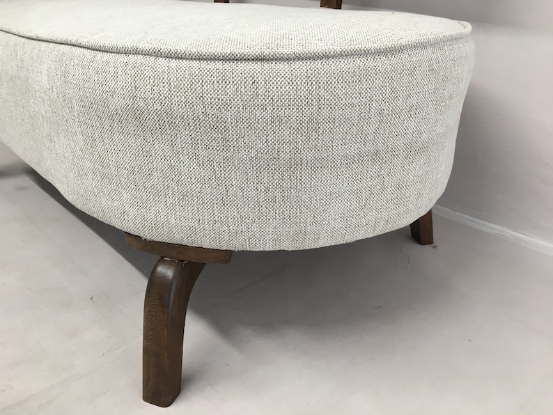 May include: A curved bench featuring light grey, textured fabric upholstery with a visible weave pattern. The seat is supported by dark brown wooden legs that curve outwards, providing a stable base for the rounded, upholstered furniture piece.