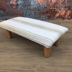 May include: A small, rectangular footstool with a white fabric top featuring beige stripes. The stool has four short, light brown wooden legs. The design is simple and the color palette is neutral.