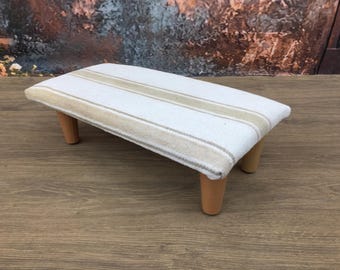 May include: A small, rectangular footstool with a white fabric top featuring beige stripes. The stool has four short, light brown wooden legs. The design is simple and the color palette is neutral.