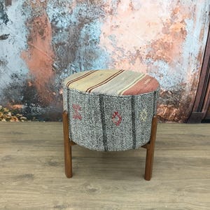 Puede incluir: Un puf redondo de color gris y beige con estampado y patas de madera. El puf está sobre un suelo de madera.