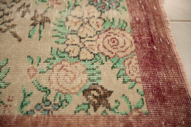 May include: Close-up of a beige rug with a floral pattern. The rug has a red border and is woven with a variety of colors, including green, pink, and blue.
