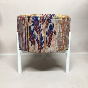 May include: A round, white-legged ottoman with a colorful floral patterned fabric cover.