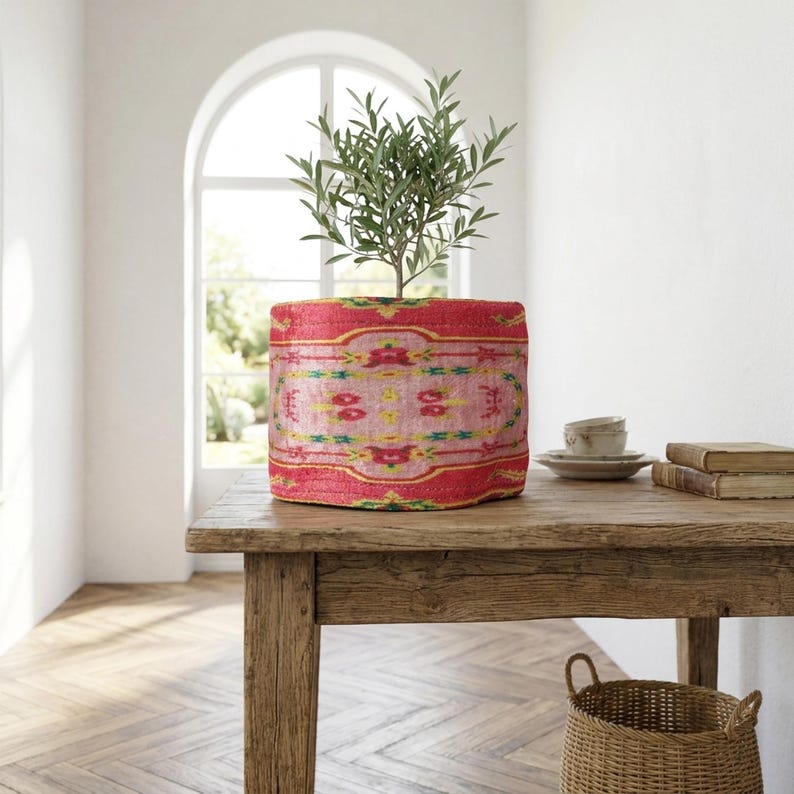 A potted plant with green leaves sits on a wooden table, accompanied by a basket and a stack of books. The room has a white wall and a window, creating a serene and inviting atmosphere.