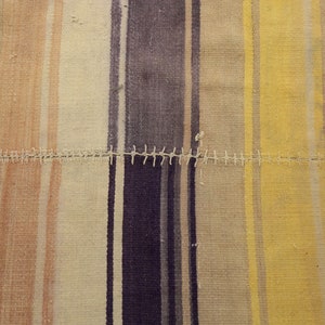 May include: Close-up of a patchwork textile with vertical stripes in shades of beige, yellow, purple, and peach. The stripes are sewn together with white thread. The textile has a rustic, handmade appearance.