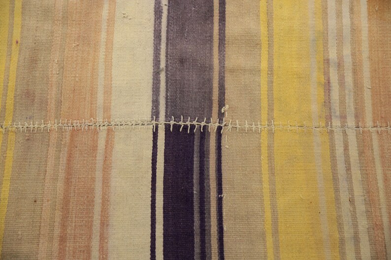 May include: Close-up of a patchwork textile with vertical stripes in shades of beige, yellow, purple, and peach. The stripes are sewn together with white thread. The textile has a rustic, handmade appearance.