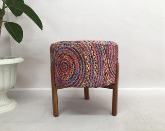 Puede incluir: Un puf redondo y colorido con un patrón y un marco de madera. El puf tiene un patrón rosa, rojo, azul y verde.