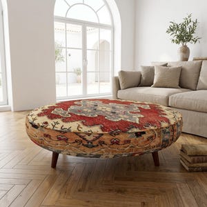 Könnte beinhalten: Ein ovaler Hocker mit einem Teppichmuster in Rot, Beige und Grau. Der Hocker hat kurze, dunkle Holzbeine und steht in einem Wohnzimmer mit einem beigen Sofa und Parkettboden. Das Teppichmuster ist detailliert und aufwendig.