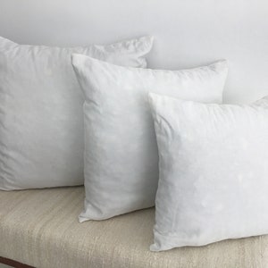 May include: Three white pillow covers of different sizes, arranged on a light brown textured surface.