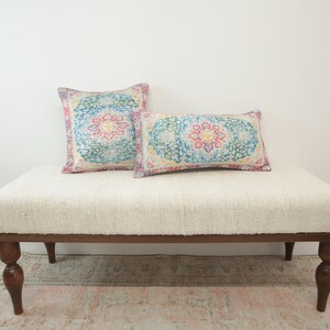 May include: A white upholstered bench with wooden legs and two patterned pillows. The pillows have a colorful floral design with shades of pink, blue, yellow, and green.