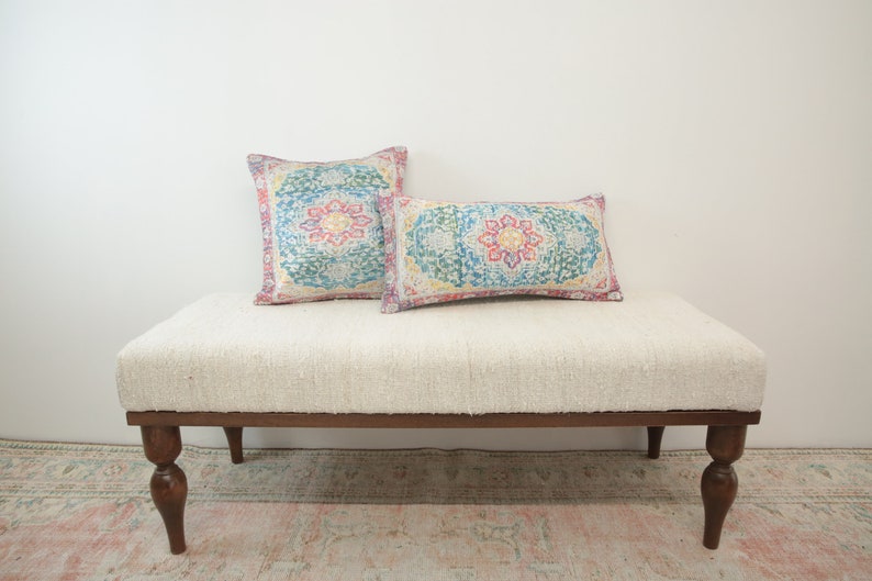 May include: A white upholstered bench with wooden legs and two patterned pillows. The pillows have a colorful floral design with shades of pink, blue, yellow, and green.
