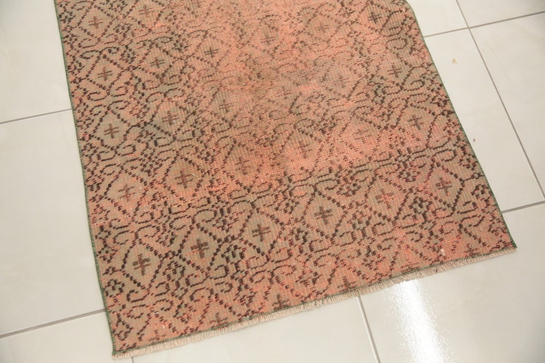 May include: A vintage rug with a geometric pattern in shades of pink, brown, and green. The rug has a fringed edge.