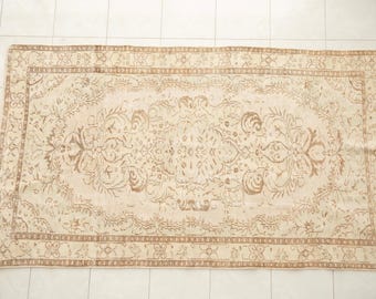 May include: A rectangular area rug featuring a light beige background with a repeating floral and vine pattern in a muted brown tone. The design includes a central medallion motif surrounded by a decorative border with geometric and organic shapes.