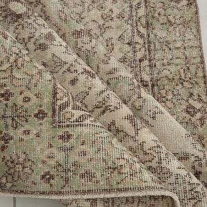 K&ouml;nnte beinhalten: Ein Vintage-Teppich mit beigefarbenem Hintergrund und einem floralen Muster in Braun- und Gr&uuml;nt&ouml;nen. Der Teppich ist in der Mitte gefaltet und zeigt die Textur des Stoffes.
