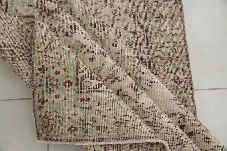 K&ouml;nnte beinhalten: Ein Vintage-Teppich mit beigefarbenem Hintergrund und einem floralen Muster in Braun- und Gr&uuml;nt&ouml;nen. Der Teppich ist in der Mitte gefaltet und zeigt die Textur des Stoffes.