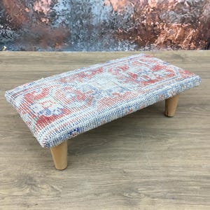 May include: A small, rectangular footstool with a patterned top in shades of red, blue, and white. The footstool has four short, light-colored wooden legs. The overall design has a vintage or antique aesthetic.