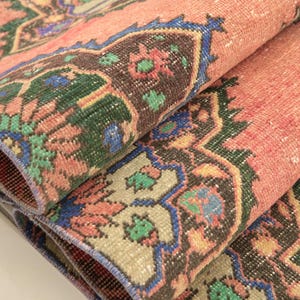 May include: A folded area rug featuring a salmon pink background with intricate geometric and floral patterns in shades of dark brown, forest green, and royal blue. The textile displays a repeating medallion design with detailed borders and edges.