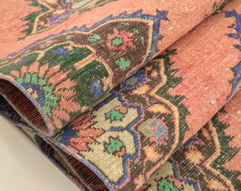 May include: A folded area rug featuring a salmon pink background with intricate geometric and floral patterns in shades of dark brown, forest green, and royal blue. The textile displays a repeating medallion design with detailed borders and edges.