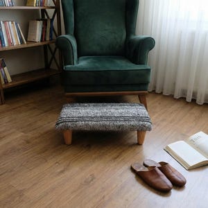 May include: A dark green armchair with a matching footstool. The footstool has a grey and black striped pattern and wooden legs. Brown leather slippers and an open book are on the wooden floor.