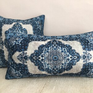 Op de afbeelding: Twee decoratieve rechthoekige kussens met een blauw en wit patroon. De kussens hebben een ingewikkeld, sierlijk patroon dat doet denken aan een traditioneel tapijt. Het kleurenpalet omvat tinten blauw, wit en grijs.