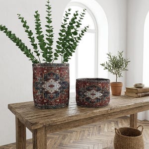 May include: Two cylindrical planters with a vintage rug design in red, blue, and cream sit on a rustic wooden table. One planter holds tall green eucalyptus branches. A smaller planter is next to it. The scene is set in a bright room.