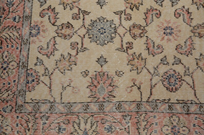 May include: A close-up of a beige rug with a floral pattern. The rug has a light brown border and a light pink center. The rug is made of a soft, woven material.