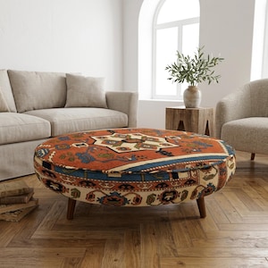 Könnte beinhalten: Ein ovaler Hocker mit einem gemusterten Teppichdesign in Rost-, Blau- und Cremetönen. Der Hocker hat kurze, dunkle Holzbeine und steht in einem Wohnzimmer mit Sofa und Sessel.