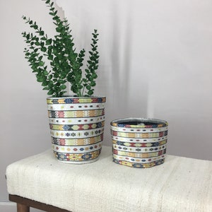 Könnte beinhalten: Zwei zylindrische geflochtene Körbe mit einem farbenfrohen geometrischen Muster in Rot-, Gelb-, Blau- und Grüntönen. Die Körbe stehen auf einer weißen Bank mit Holzbeinen.