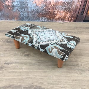 May include: A small, rectangular footstool with a patterned top and wooden legs. The top features a geometric design in shades of black, blue, and white. The legs are short and cylindrical, providing a stable base for the stool.