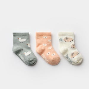 May include: Three pairs of baby socks in soft pastel colors. One pair is gray with white duck illustrations, another is peach with white flower designs, and the third is off-white with sheep. The word "kumori" is printed on each sock.