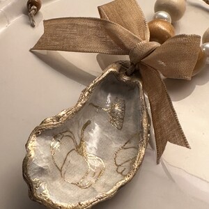 May include: A white oyster shell with gold leaf accents and a floral design. The shell is attached to a brown ribbon and a beaded necklace with wooden and pearl beads.