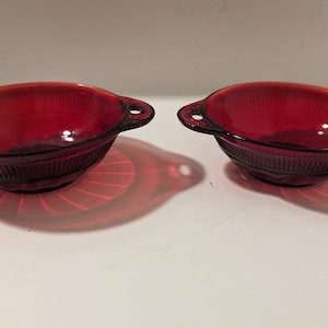 May include: Two red glass bowls with handles. The bowls have a ribbed design and are made of ruby red glass.
