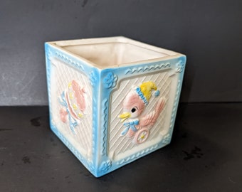 May include: A white ceramic planter shaped like a cube with blue and white trim. The planter has a design of a pink bird with a yellow hat and a pink baby carriage on the front.