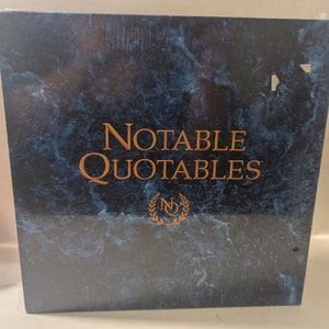 May include: A blue book with a gold marble pattern and the text "Notable Quotables" in gold lettering. A gold laurel wreath with the letter "N" inside is below the text.