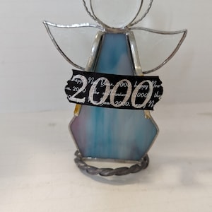 Op de afbeelding: Een glas-in-lood engelfiguur met een blauw en roze lichaam en een halo. De engel houdt een bord vast met de tekst "Happy New Year 2000".