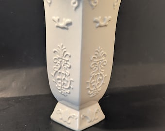 May include: A white ceramic vase with an ornate design. The vase has a scalloped rim and a hexagonal base.