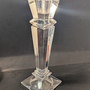 May include: A clear glass candle holder with a square base and a tall, rectangular body. The holder has a geometric design with multiple facets.