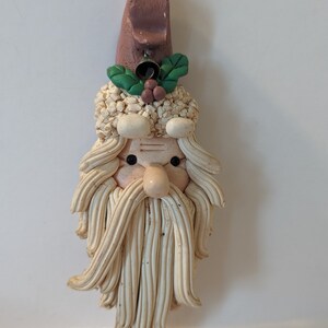 May include: A white ceramic Santa Claus ornament with a brown hat, green holly, and a small bell. The ornament has a white beard and a detailed face.