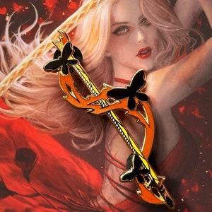Puede incluir: Un pin de esmalte dorado y naranja con un arco y una flecha estilizados con detalles de mariposas negras. El pin está colocado sobre un fondo de una mujer con el pelo largo y rubio que lleva un vestido rojo.