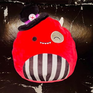 May include: A red plush toy character with a black and white striped lower section, a black top hat with a purple band and a white cross, and a winking eye. It has a small, smiling mouth and a white horn.