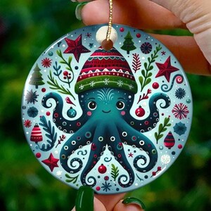 May include: A round Christmas ornament featuring a teal octopus wearing a red and green hat. The ornament has a light blue background with red stars, green trees, and other festive designs. A gold string is attached for hanging.